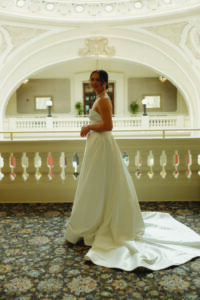 Bride in strapless white wedding dress smiles indoors by ornate railing, with decorative ceiling and arched window behind her.