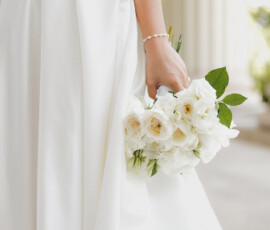 Bride in white dress holding bouquet, lower body visible, with white columns and greenery in elegant Mobile Alabama setting.