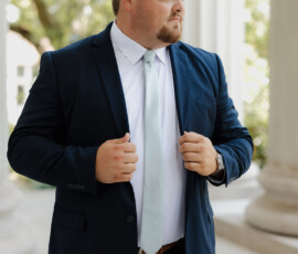 Man in navy suit and light tie adjusts his jacket by white columns, before a Mobile Alabama wedding.