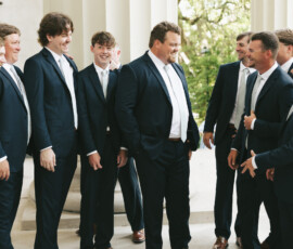 Group of groomsmen in navy suits laughing together outside a columned wedding venue before the ceremony.