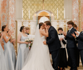 Bride and groom sharing their first kiss at the altar as wedding party members applaud.