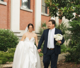 Bride in white gown and groom in navy suit stroll hand-in-hand by brick building and greenery at Battle House Mobile wedding.