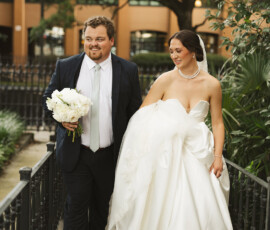 Bride and groom walk hand in hand on a bridge at their Mobile, Alabama wedding, smiling with white flowers and scenic trees behind.