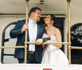 Newlywed couple smiling at each other while riding on the back platform of a vintage white trolley.