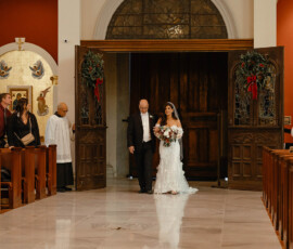 Bride in white dress walks with older man down cathedral aisle; festive doors with greenery and red ribbons in downtown Mobile.