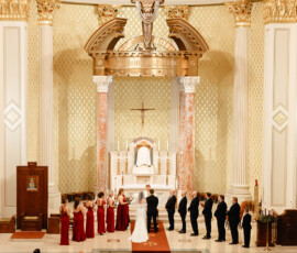 Bride and groom at altar in ornate Mobile, Alabama church, with bridesmaids in red and groomsmen in black as guests look on.
