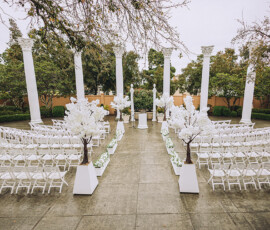 Elegant outdoor New Orleans wedding with white chairs, decorative trees, and columns lining the aisle to a greenery-framed altar.