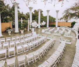 Outdoor wedding venue with semicircle rows of white chairs, elegant floral decor, white columns, and lush green backdrop.