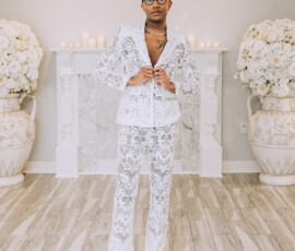 Person in a white lace suit and boots stands in a bright room with floral arrangements, candles, and wood floors—chic wedding inspiration.