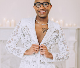 Smiling person in glasses and a white lace suit reveals chest tattoo and necklace by lit candles at a Louisiana wedding.