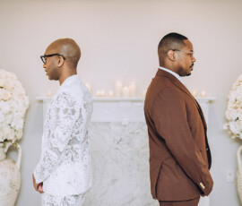 Two men in white and brown suits stand back to back by a decorated fireplace at a Louisiana wedding.