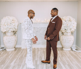 Two men smile at each other in a bright room with white floral decor; one in a white lace suit, the other in a brown suit.