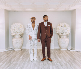 Two men in white floral and brown suits hold hands in a bright room with flowers—stylish Gulf South wedding inspiration.