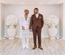 Two men holding hands, smiling at each other in a bright room with white flowers at a joyful Louisiana wedding.