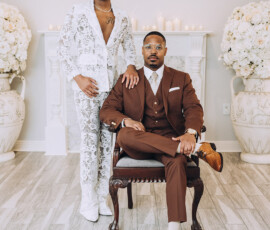 Two men, one in a white lace suit and one in brown, pose among white flowers and candles in a bright, romantic wedding scene.