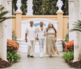 Three people holding hands walk among flowers and palms, framed by marble columns at an elegant New Orleans wedding.
