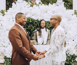Two grooms hold hands and smile at their Louisiana outdoor wedding, with an officiant by a white floral arch.