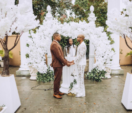 Two people in formal suits hold hands before a floral arch and officiant, amid white blossoms at a Gulf South outdoor wedding.