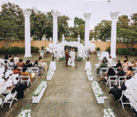 Outdoor wedding with guests, white flowers, and tall columns. Couple stands at altar before officiant. Timeless inspiration.