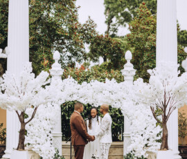 Outdoor wedding ceremony with two people holding hands, an officiant, white flowering trees, and tall white columns.