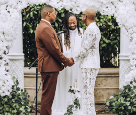 Two grooms hold hands under a white floral arch with a smiling officiant during an outdoor New Orleans wedding ceremony.