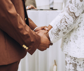 Couple holding hands at a Louisiana wedding, wearing a brown suit and white lace outfit, standing close together.
