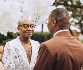 Two people face each other at an outdoor New Orleans wedding, one in a white lace suit and the other in brown, with flowers and a crowd.
