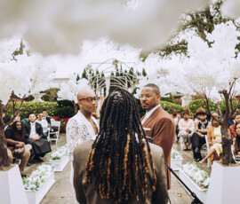 Two grooms face each other at an outdoor wedding with guests, white flowers, and the officiant standing between them.