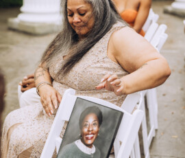 Woman in beige lace dress holds framed photo at outdoor Louisiana wedding, with other guests in the background.