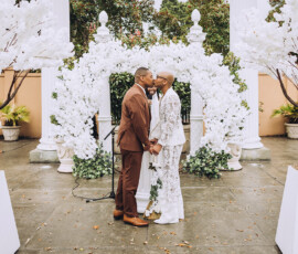 Couple holding hands and smiling at each other during an outdoor New Orleans wedding, surrounded by white flowers and trees.