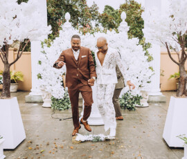 Two grooms in brown and white suits celebrate, holding hands at an outdoor wedding with white flowers and decorative trees.