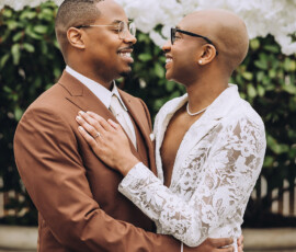 Two people embrace and smile, one in a brown suit and one in white lace with an engagement ring, against a dreamy floral backdrop.