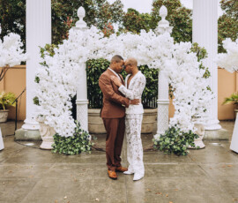 Couple embraces by white floral arches at an outdoor wedding, surrounded by greenery and decorative white trees.