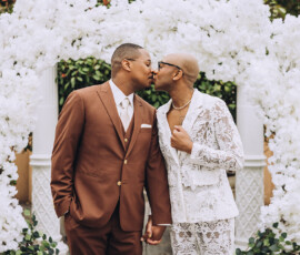Two men kiss, holding hands under a white floral arch at a Gulf South wedding; brown suit, white lace suit, greenery behind.