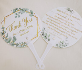 Two paper wedding fans with green foliage and gold accents, featuring thank you messages, on light fabric.
