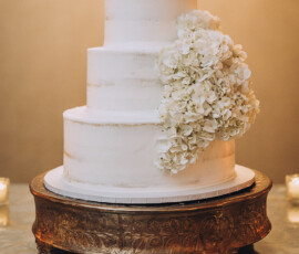 Three-tiered white wedding cake with rustic frosting and white hydrangeas on a gold stand—elegant Gulf South wedding inspiration.