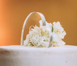 White frosted cake with white flowers and an arched acrylic topper, set against a warm, blurred background.
