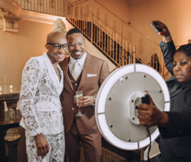Two men in formal wear smile for a photo as a woman takes it with a phone and ring light at an elegant wedding reception.