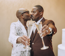 Two men in formalwear kiss near a white wedding cake, holding champagne and celebrating their wedding ceremony.