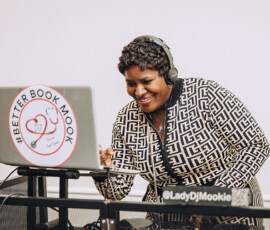 Smiling DJ in black-and-white outfit mixes music at a laptop with #BetterBookMook sticker at Gulf South wedding DJ booth.