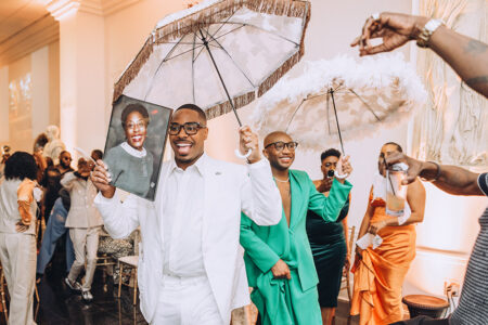 Newlyweds dancing through their reception entrance holding decorative umbrellas during a lively second line celebration.