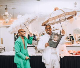 Two men in bright suits hold decorated parasols at a lively New Orleans wedding with chandeliers and guests in the background.