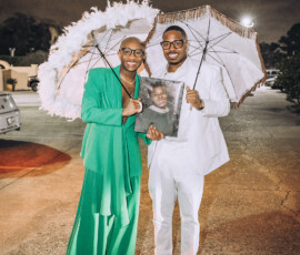 Two people in formal attire, holding ornate umbrellas and a portrait, smile outdoors at night during a joyful Louisiana wedding.