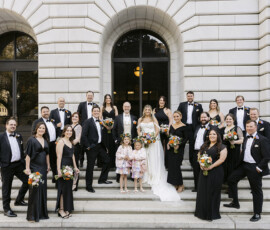Wedding party portrait on stone steps outside a historic building with attendants in black formal attire holding bouquets.