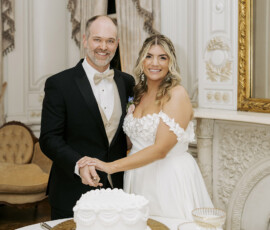 Couple cutting a white wedding cake inside an elegant historic venue with ornate molding and gold mirror.