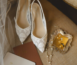 White lace bridal shoes, gold rings, pearl necklace, veil, and vow books on a beige chair for a classic Louisiana wedding.