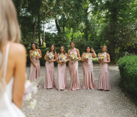 Bride and six bridesmaids in blush pink dresses with bouquets on a gravel path amid greenery—Louisiana wedding inspiration.