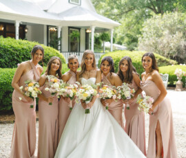 Bride in white gown with six bridesmaids in blush pink, smiling with bouquets outdoors by a white house and greenery.