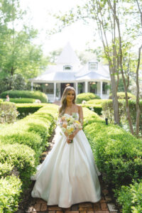 Bride holding pastel bouquet while standing on a garden pathway with historic white house in the background.