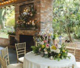Round table with floral centerpiece and gold chairs beside a brick fireplace with flowers and candles for garden wedding inspo.